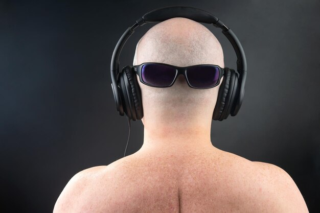 Foto indietro di un uomo nudo che ascolta musica con cuffie e occhiali da sole sulla parte posteriore della testa su uno sfondo scuro