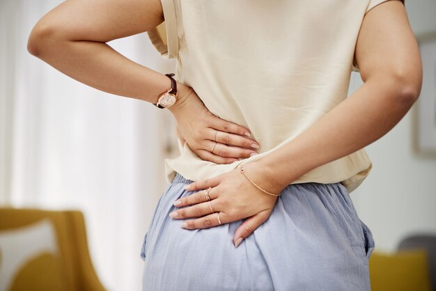 自宅で不快感や関節炎に問題があるリビングルームにいる腰痛の手と女性 腰痛脊椎と、骨粗鬆症や線維筋痛症でラウンジで関節や姿勢がけいれんする人