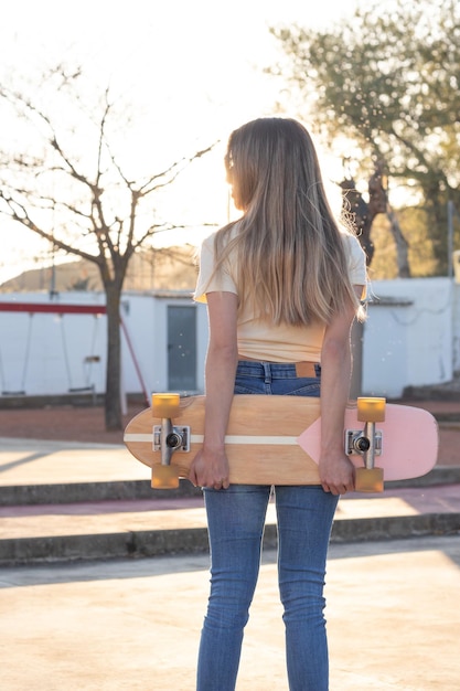 Ritratto posteriore di una giovane ragazza bionda esile che tiene uno skateboard vintage naturale dietro la schiena