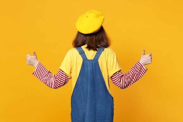 Back rear view of bright girl teenager in french beret, denim sundress showing thumbs up isolated on yellow wall background in studio. people sincere emotions, lifestyle concept. mock up copy space