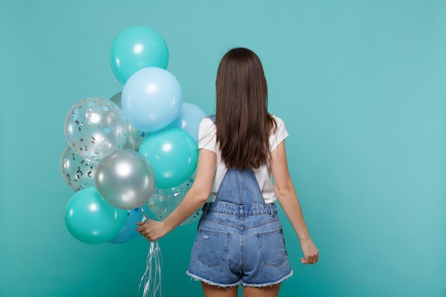 Back rear view of slim young brunette woman in denim clothes celebrating and holding colorful air balloons isolated on blue turquoise wall background. Birthday holiday party, people emotions concept.