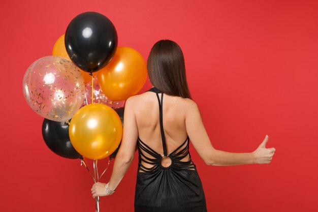 Back rear view of young girl in little black dress celebrating showing thumb up hold air balloons isolated on red background. St. Valentine's Day Happy New Year, birthday mockup holiday party concept.