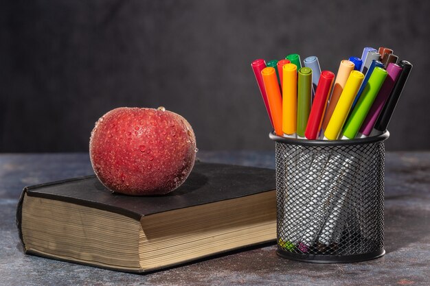 Foto di nuovo a scuola. apple in gocce d'acqua che giace sul libro, accanto a pennarelli multicolori in piedi nel supporto