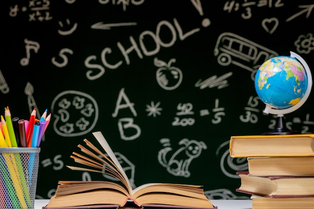 Back to school background with books, pencils and globe on white table on a green blackboard background.