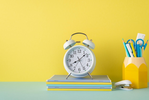 back to school concept photo of school accessories on blue desk alarm clock over stack of copybooks mini stapler and stand for pens on yellow wall background