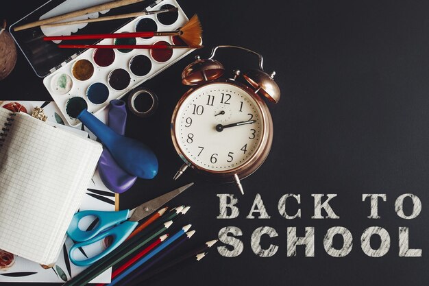 Foto ritorno a scuola concetto testo gesso bianco su lavagna nera matite colorate vernici pennelli forbici e taccuino con vecchio orologio sullo spazio per l'educazione piatta laica del testo