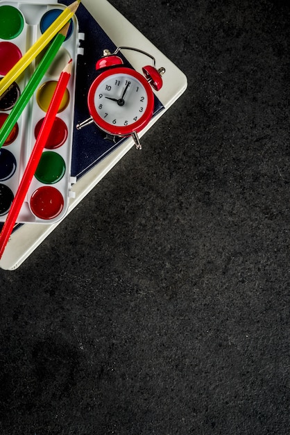 Di nuovo al concetto della scuola con il fondo della lavagna delle matite di colore della sveglia dei libri