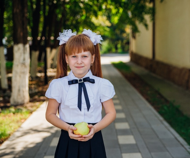 Di nuovo a scuola. una graziosa scolaretta è in piedi nel parco o nel cortile della scuola e tiene in mano una mela verde. pasti scolastici adeguati per il pranzo. una bambina sta andando in prima elementare.