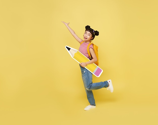 Back to school. Happy cute asian child with big backpack holding pencil on yellow wall. education concept.