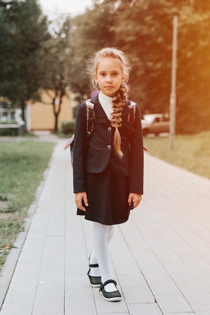 Ritorno a scuola piccolo bambino felice studentessa di otto anni in uniforme di moda con zaino e acconciatura voluminosa lunga treccia pronta per andare in seconda elementare primo giorno alla scuola primaria flare