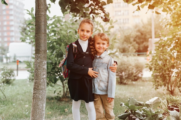 Back to school little happy kid pupil schoolgirl eight years\
old in fashion uniform with backpack and her preschool brother boy\
hug together ready going second grade first day primary school\
flare