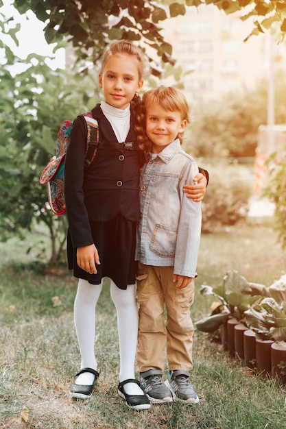 Back to school little happy kid pupil schoolgirl eight years old in fashion uniform with backpack and her preschool brother boy hug together ready going second grade first day primary school flare