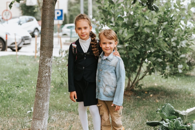 Back to school little happy kid pupil schoolgirl eight years\
old in fashion uniform with backpack and her preschool brother boy\
hug together ready going second grade first day primary school