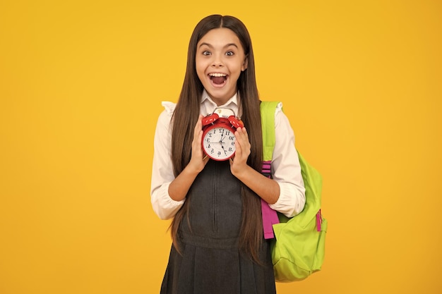 Photo back to school teenager school girl with backpack hold clock alarm time to learn school children on isolated yellow background happy girl face positive and smiling emotions