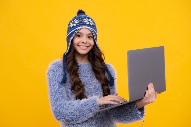 Back to school teenager schoolgirl with warm hat and sweater hold laptop on yellow isolated studio background winter school happy face positive and smiling emotions of teenager girl