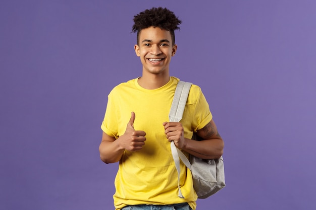 Back to school, university concept. Portrait of cheerful handsome male student describe his summer vacation to classmates, show recommendation, thumbs-up approval, hold backpack