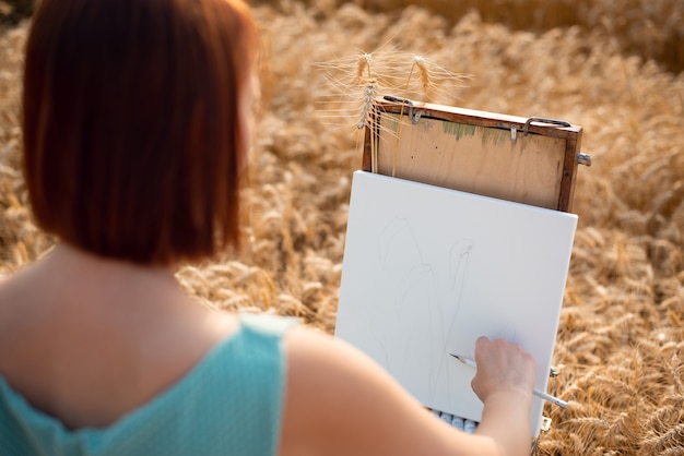 熟した穀物の分野で白い帆布に絵を描き始めている女の子の画家のバックショット