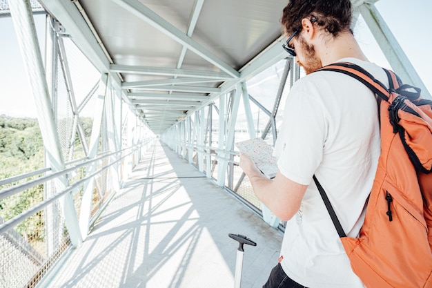 Back shot of a traveler checking a map while carrying his\
luggage and bags at the airport or bus station during a travel.\
hipster modern traveler, sunny day, copy space.