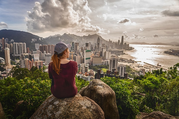 Back side of asia woman traveler sitting after climbing the peak of hong kong mountain and looking hong kong and kowlloon cityscape when sunset time, adventure