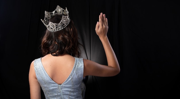 Back Side view Portrait of Miss Pageant Beauty Contest in sequin Evening Ball Gown long dress sparkle light Diamond Crown, Asian Woman fashion make up black hair style, studio lighting dark background