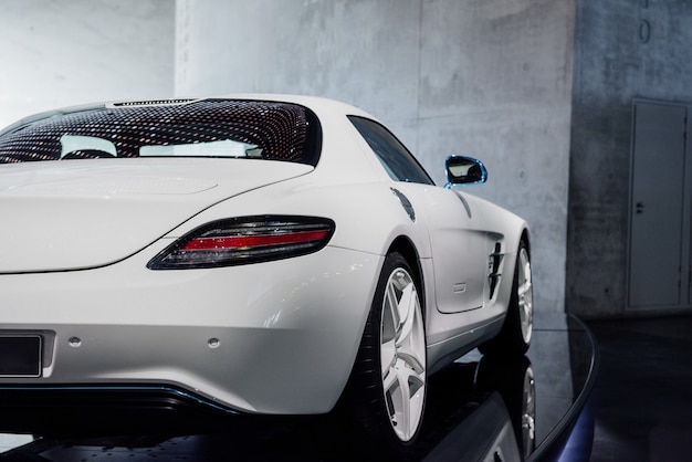 Back, side view of white cool sport car with right diode backlight, blue mirror with turn signal, trunk, light alloy wheels and low-profile tires, reflection of lights on window near the grey wall.