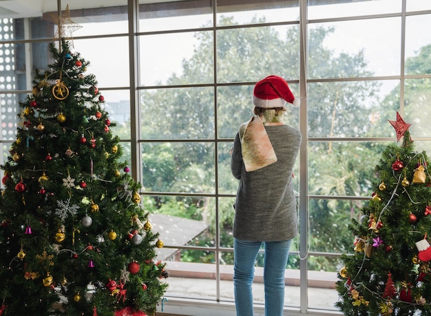 Back of standing Caucasian senior woman with Santa hat alone at home with decorated Christmas tree looking outside window on Christmas and new year celebration festival. lonely holidays concept.