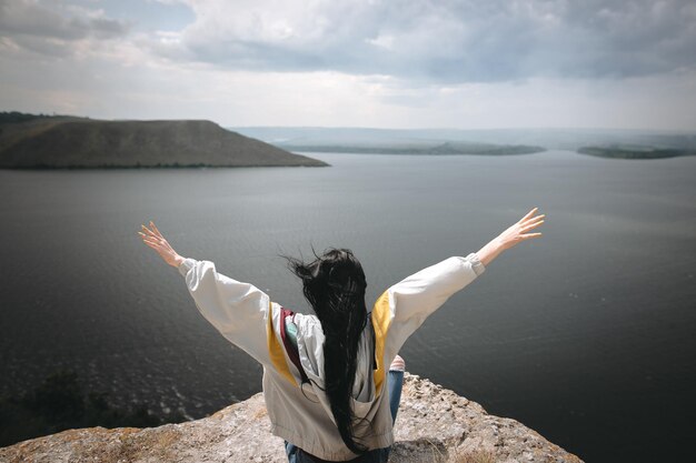 Back of stylish hipster girl sitting on top of rock mountain with beautiful view on river Young tourist woman with windy hair relaxing on cliff Travel and wanderlust Copy space