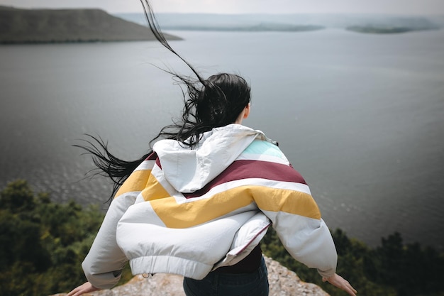 Back of stylish hipster girl on top of rock mountain with beautiful view on river Young tourist woman with windy hair relaxing on cliff Atmospheric moment Copy space