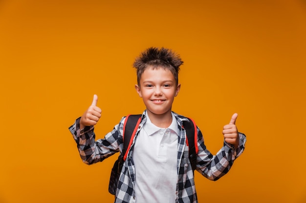 Снова в школу забавный мальчик с рюкзаком в рубашке на желтом фоне показывает суперкласс и улыбается смотрит в камеру для копирования образование
