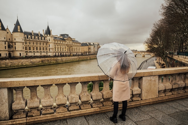 Back of traveler young girl walking in Paris in the rain day and holds transparent umbrella in his hand.