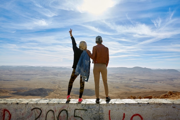 The back of two women, stand with their hands up overlooking the mountains.