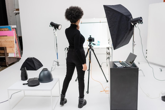 back view of african american content manager in black clothes standing near laptop and digital