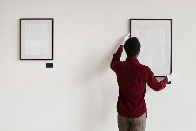 Photo back view at african-american man hanging blank frames on wall while planning art gallery or exhibition,