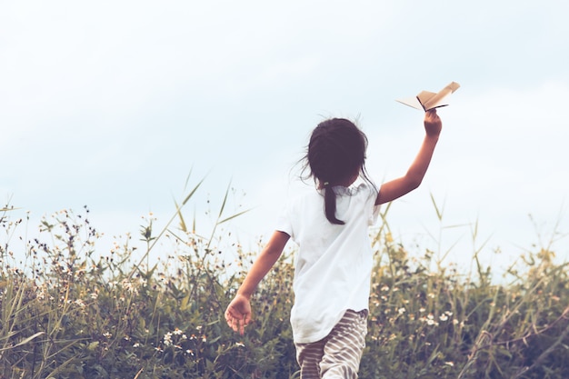 ビンテージ色のトーンで牧草地でおもちゃの紙飛行機を遊んでいるアジアの子供の背面図