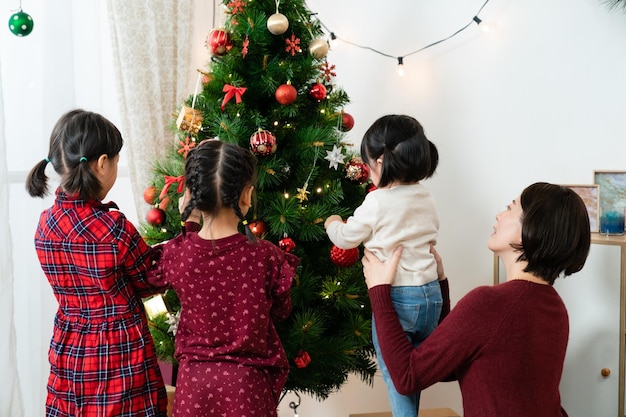 自宅の居間で兄弟姉妹と一緒にクリスマスツリーに飾りをつけている末娘を支えるアジア人の母親の後ろ姿