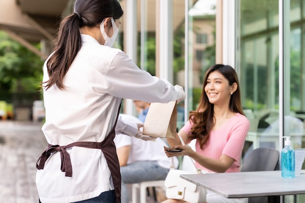 フェイスマスク付きのアジアのウェイトレスの背面図は、魅力的な女性の女性の顧客にフードバッグを取り出す注文を与えます。コロナウイルスのパンデミック後の新しい通常のテイクアウトまたはテイクアウトのフードサービスの概念。