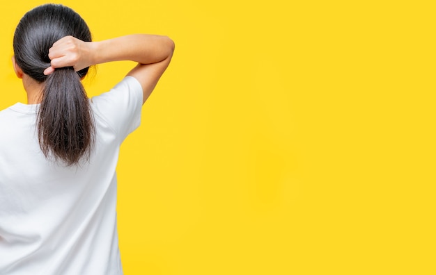 Back view of Asian woman holding damaged hair on yellow background with copy space. Split ends hair problem in woman. Dry and brittle black long hair needs shampoo and conditioner for spa treatment.