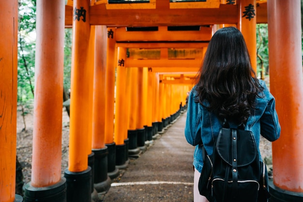 Vista posteriore della donna asiatica con bei capelli lunghi neri lisci in piedi in rosso torii. stile di vita da viaggiatore con zaino e sacco a pelo in giappone. traduzione sul testo del cancello 