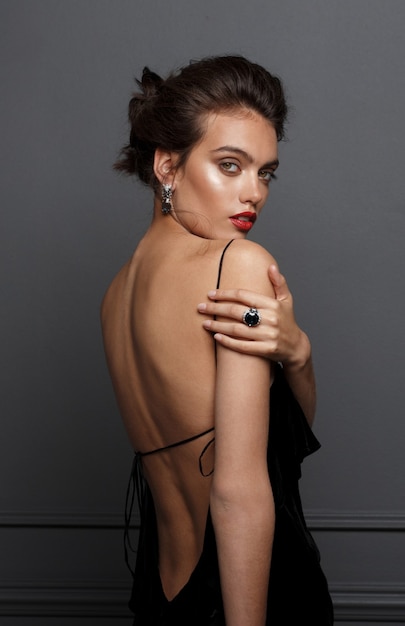 Photo back view of a attractive female model in black dress with bare shoulders, wears blue stone earrings and rings over grey dark background.