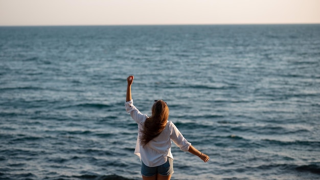 Back view of attractive woman with raised hands in a white shirt and shorts standing near the sea and enjoying the view Panoramic picture