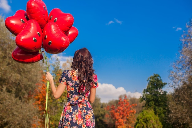 Back view of attractive young girl with red smiling balloons in hand outdoor