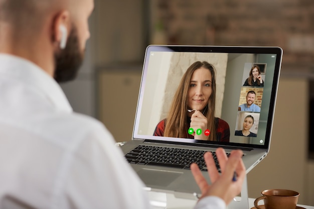 Back view of a bald male employee in earphones who is working remotely gesturing during a business video conference on a computer from home.