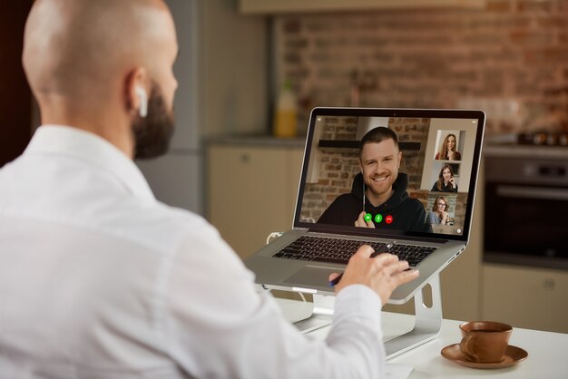 Photo back view of a bald male employee in earphones who is working remotely gesturing during a business video conference on a laptop.