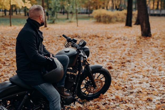 Back view of bearded male biker looks thoughtfully somewhere
into distance poses on black motorbike holds protective helmet
spends leisure time in autumnal park copy space for your
advertising