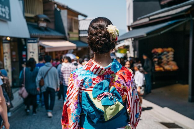 back view of beautiful Japanese lady with kimono and blue bow on the belt walking on the teeming street. Asia Japanese woman lifestyle. young girl in colorful dress walking on sunny day.