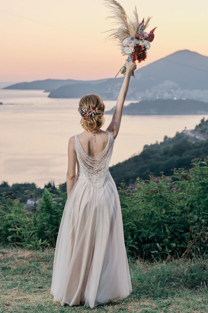 Vista posteriore della sposa in piedi sulla montagna e alzando la mano con un bouquet color up cerimonia di nozze in natura al tramonto