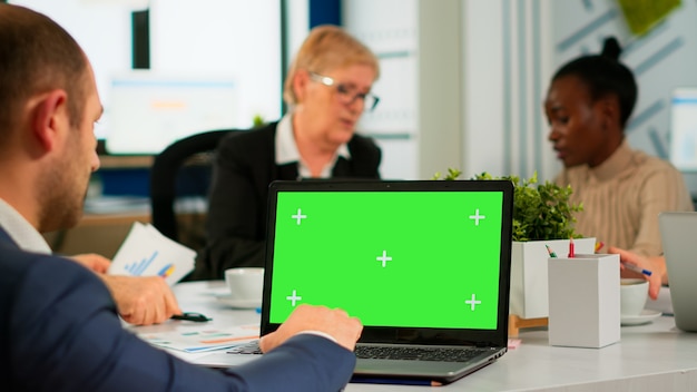 Back view of business man sitting conference desk using laptop with green screen talking with colleague while team working on background. Multiethnic coworkers planning project on chroma key display