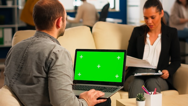 Back view of business man sitting on couch using laptop with green screen talking with colleague while team working on background. Multiethnic coworkers planning project on chroma key display