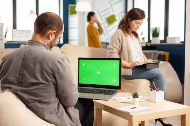 Back view of business man sitting on couch using laptop with green screen talking with colleague while team working on background