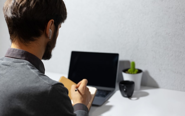 Photo back view of businessman working home, making notes in notebook, using wireless earphones, laptop, coffee mug and cactus on work desk.